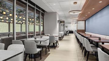 A modern restaurant with long tables and chairs, large windows, and a ceiling with recessed lighting.