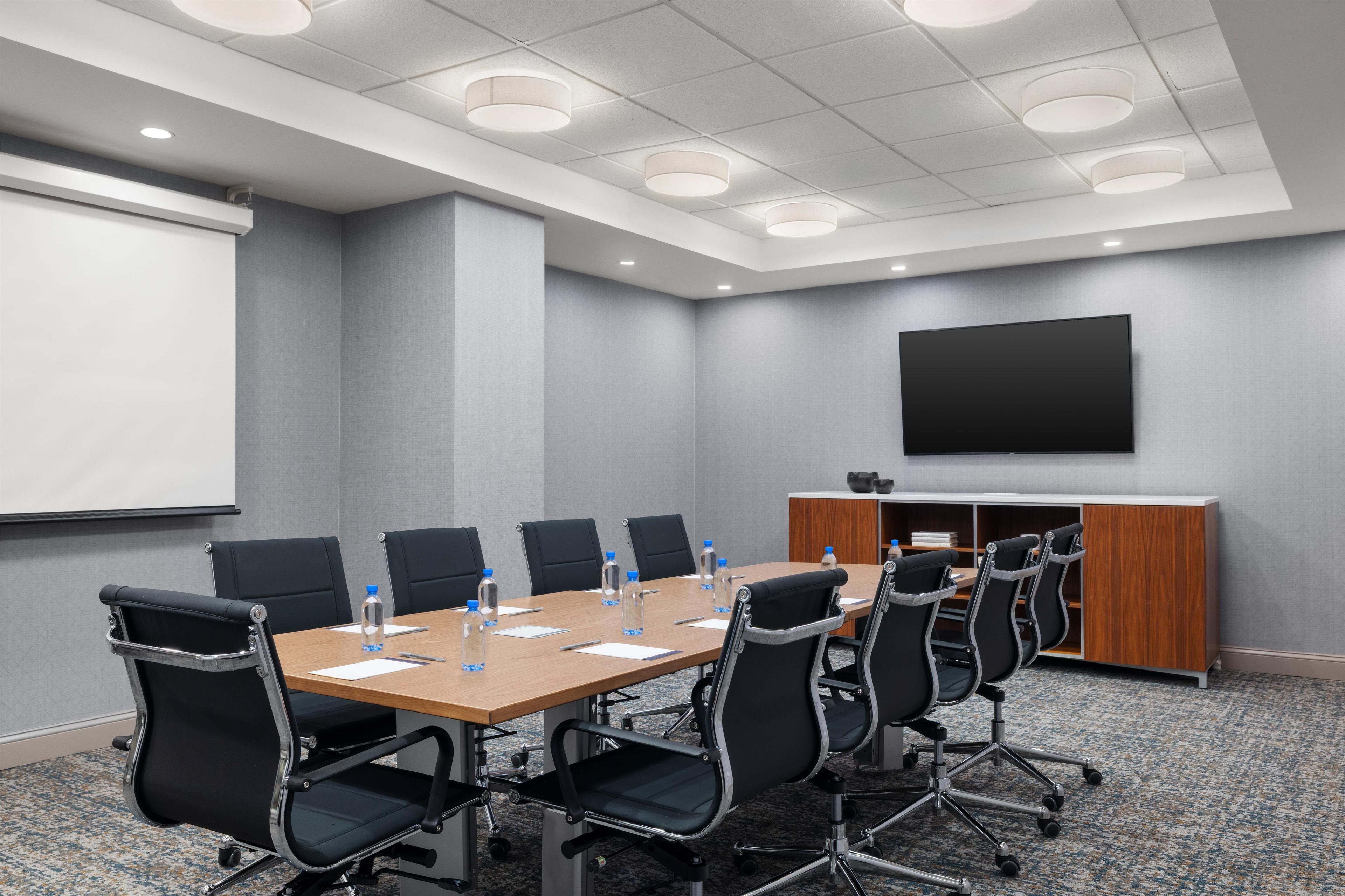 A conference room with a long table, black chairs, a whiteboard, and a flat-screen TV on the wall.