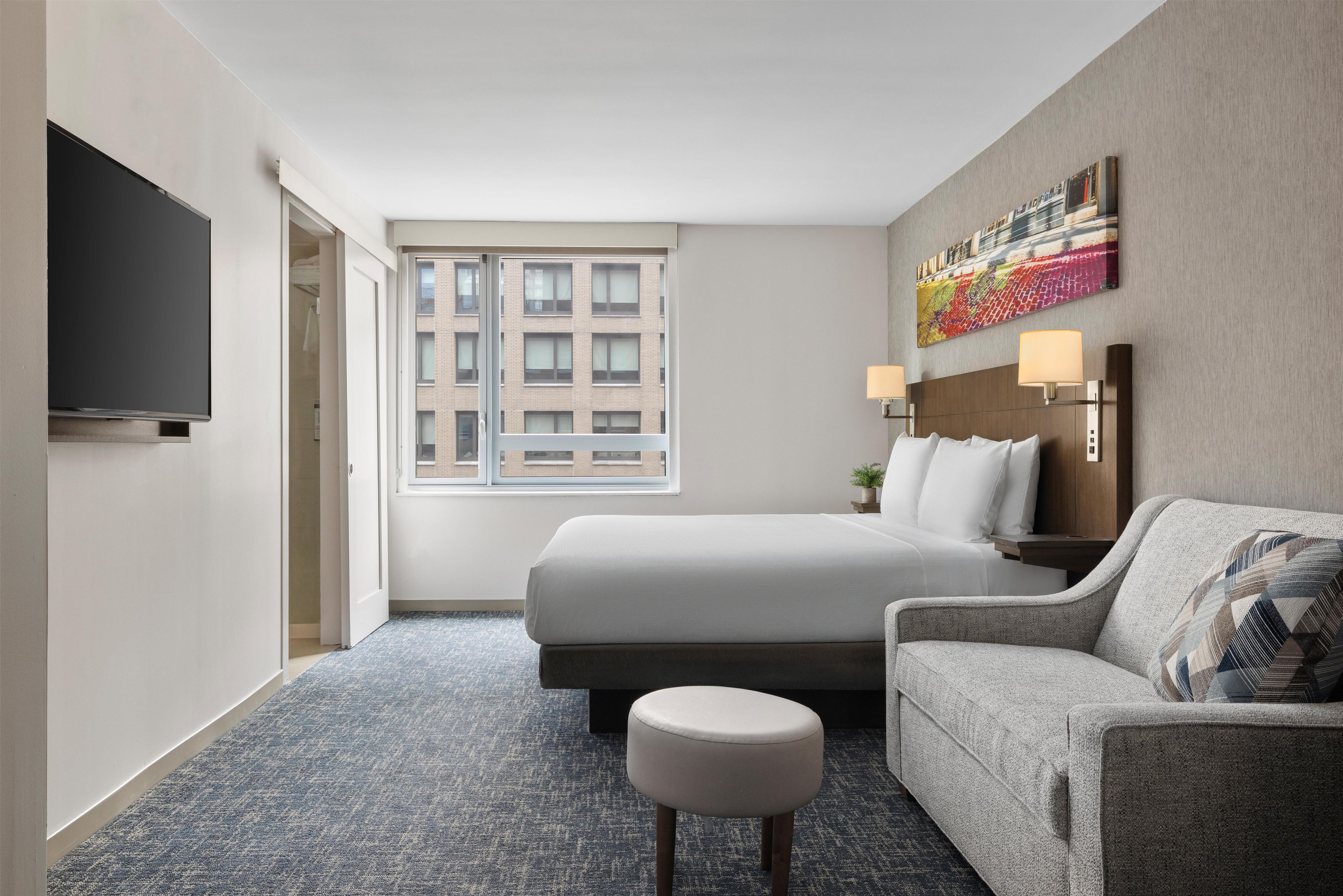 A hotel room with a bed, a sofa, a TV, a window, and a painting on the wall.