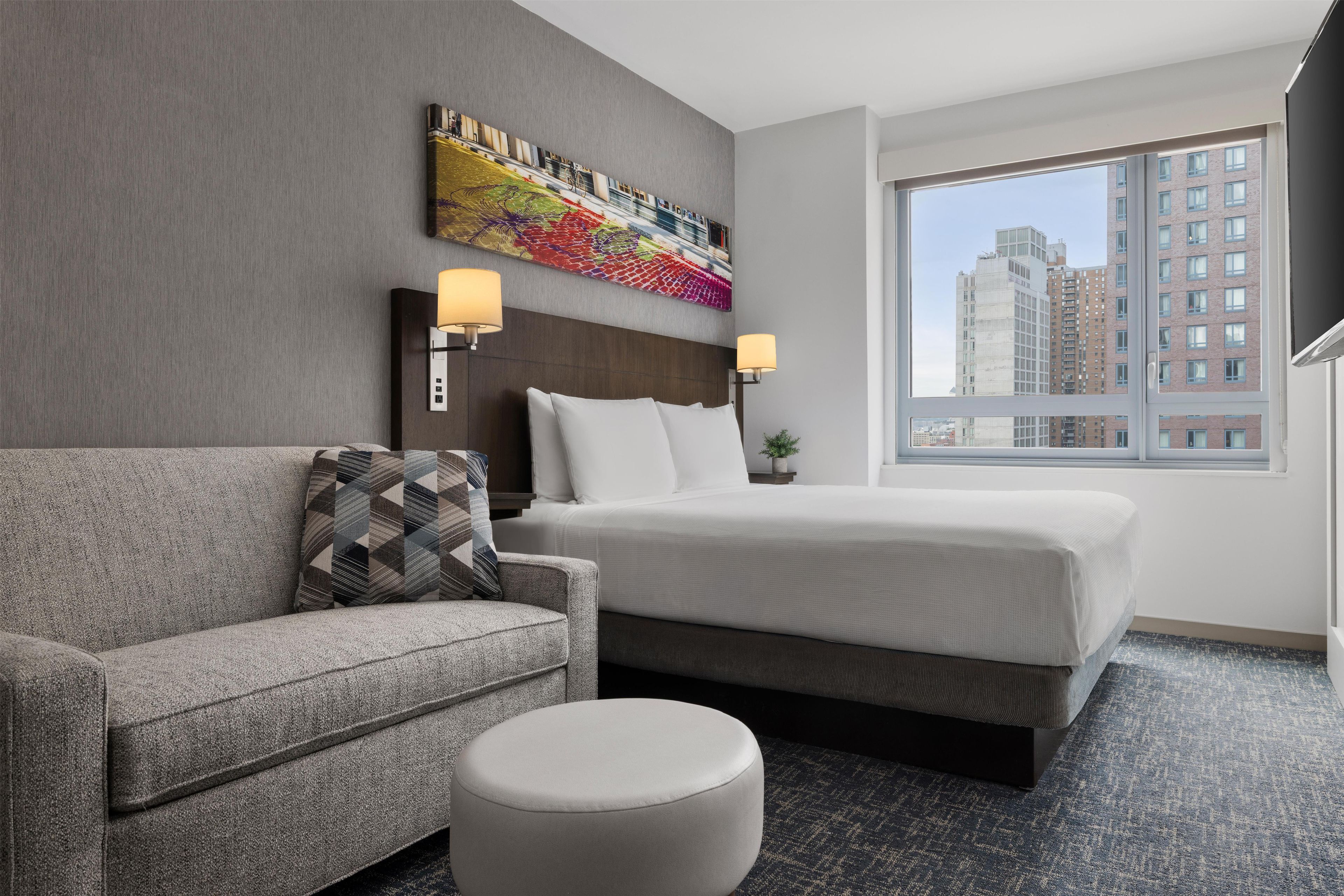 A hotel room with a bed, a sofa, a small ottoman, a nightstand with lamps, a colorful artwork on the wall, and a window with a city view.