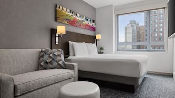 Una habitación de hotel con una cama, un sofá, un pequeño taburete, una mesita de noche con lámparas, una obra de arte colorida en la pared y una ventana con vistas a la ciudad.