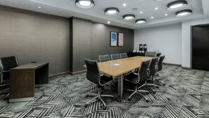Ein Konferenzraum mit einem rechteckigen Tisch, schwarzen Stühlen, einem gemusterten Teppich, deckengleichen Leuchten und gerahmten Bildern an der Wand.
