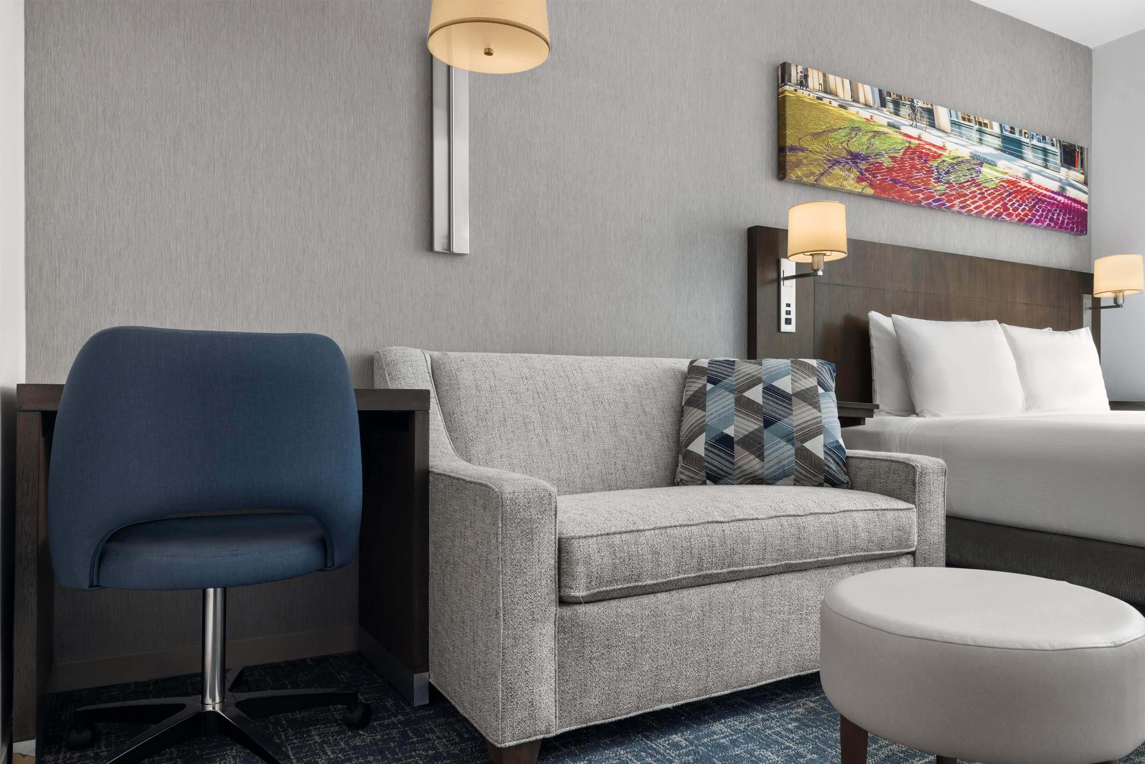 Et hotelværelse med en grå sofa, en blå lænestol, en hvid seng og et farverigt kunstværk på væggen.