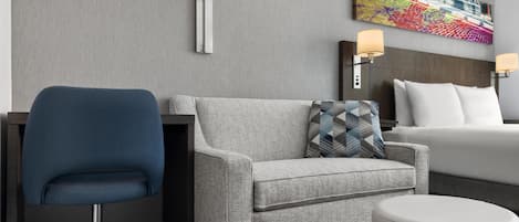 Una habitación de hotel con un sofá gris, un sillón azul, una cama blanca y una obra de arte colorida en la pared.