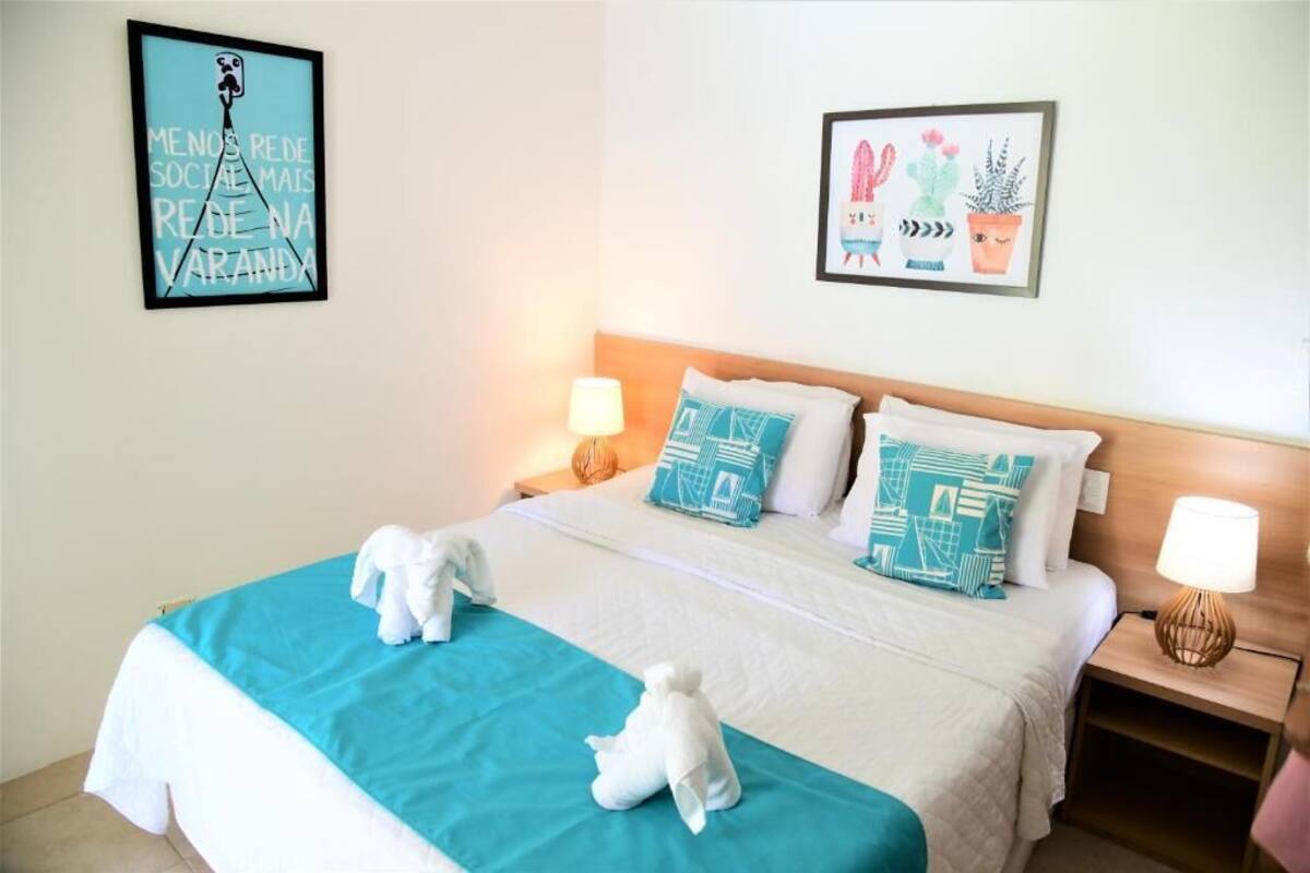 A neatly made bed with a teal bedspread and two white towels, a framed picture of plants on the wall, and a framed quote on the left.