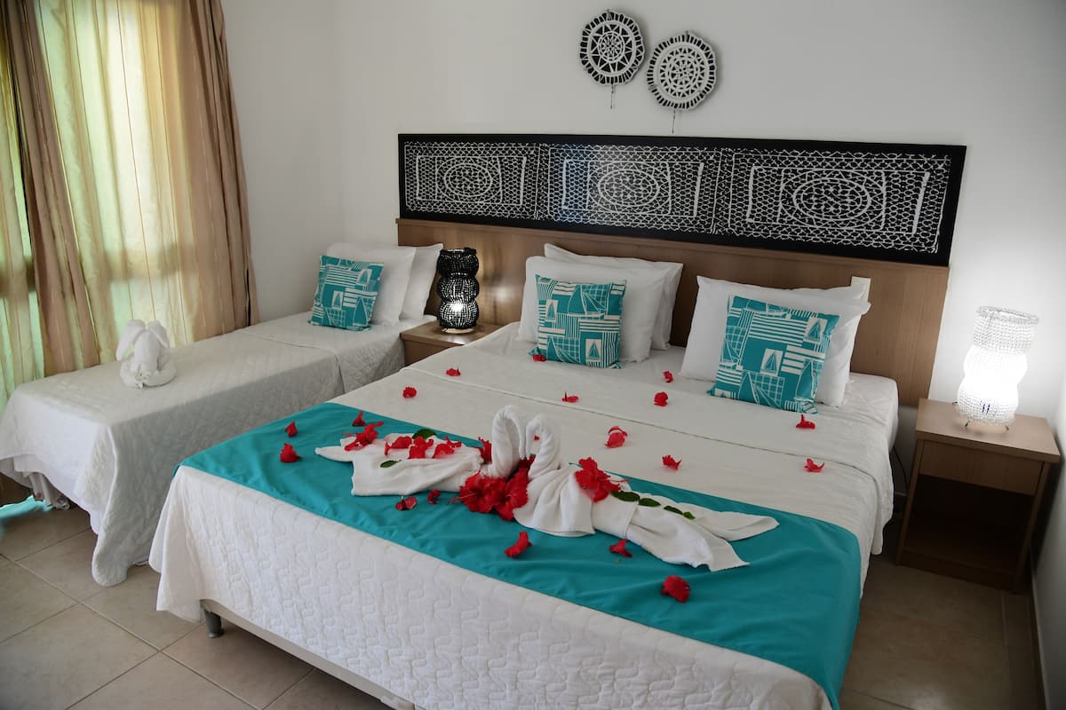 A hotel room with two beds, each with a teal bedspread, white linens, and decorative pillows. There are wall art pieces above the beds and a nightstand with a lamp.