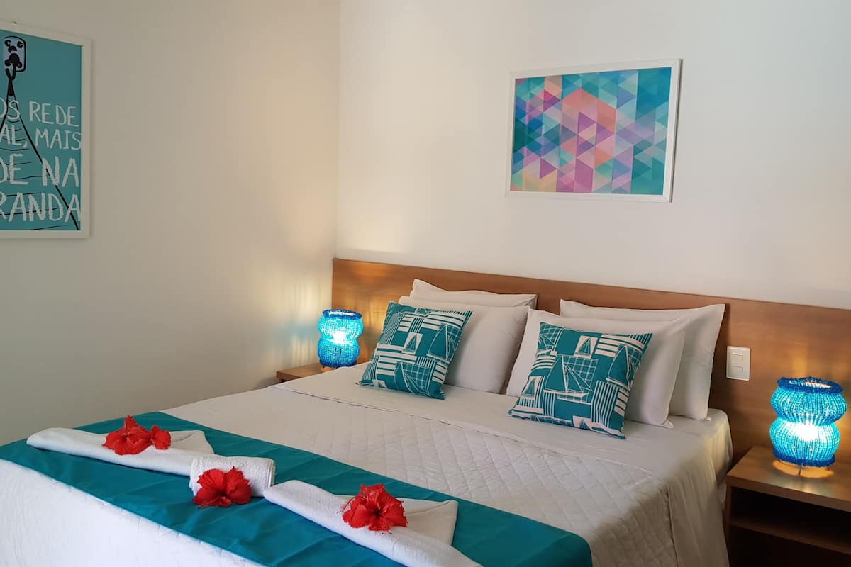 A neatly made bed with teal and white bedding, two pillows with nautical designs, and a decorative blue vase on the nightstand.