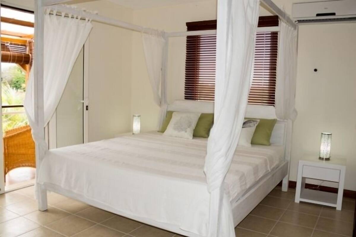 A four-poster bed with white bedding, a nightstand with a lamp, and a window with blinds.