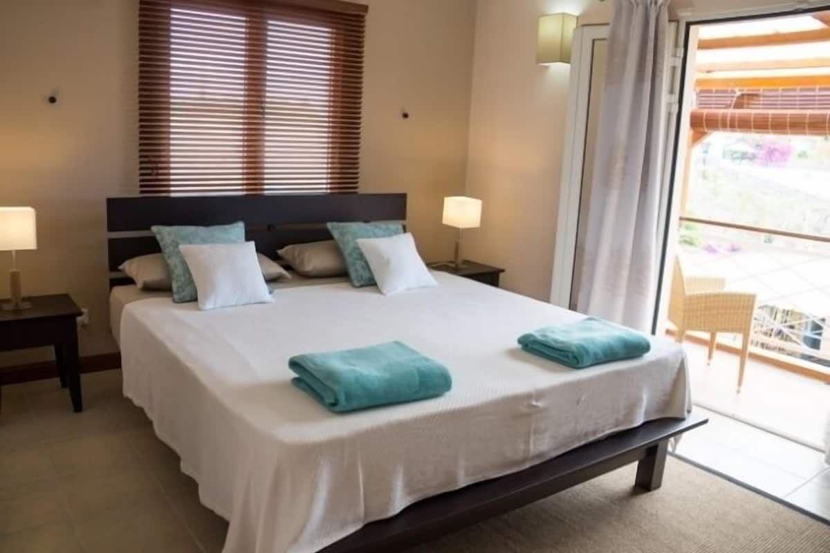 A neatly made bed with a white bedspread and two folded blue towels, a nightstand with a lamp, and a window with blinds.