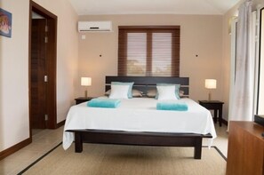 A hotel room with a large bed, two bedside tables, a window with blinds, and a door. - Villa Kreola (Grand Gaube)