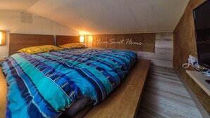 Ein Schlafzimmer mit schrägem Dach, einem Bett mit gemustertem Überwurf, zwei Wandleuchten und einem Holzkopfende mit der Aufschrift 'Home Sweet Home'.
