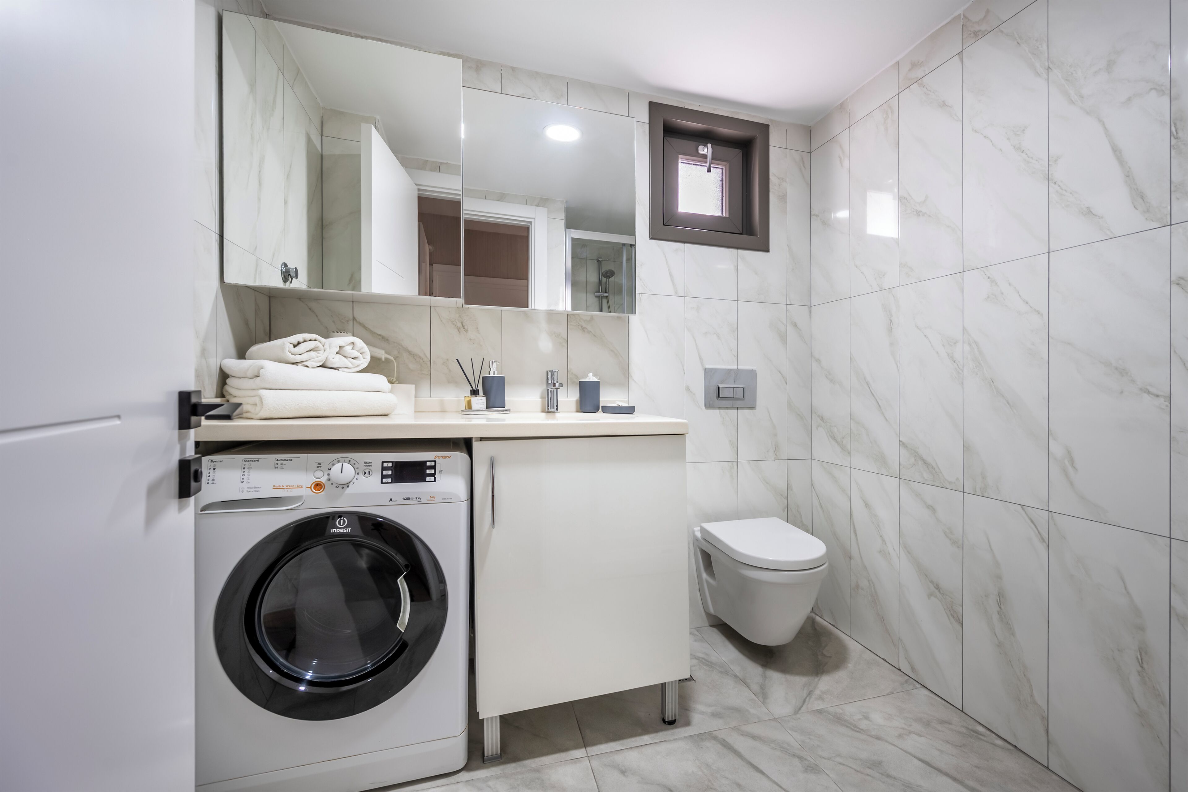 A modern bathroom with a built-in washing machine, a toilet, and a large mirror.