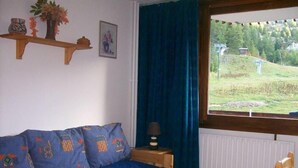 Ein Zimmer mit einem hölzernen Bett, einem blauen Sofa mit gemusterten Kissen, einem Fenster mit Blick auf eine grüne Landschaft und einem an der Wand befestigten Regal mit Dekorationen.