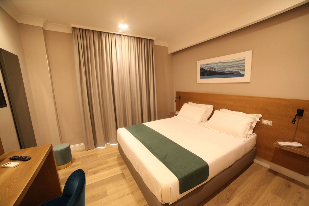 Quarto de hotel com uma cama grande, uma escrivaninha com cadeira, um pequeno banquinho, um quadro na parede e uma janela com cortinas.