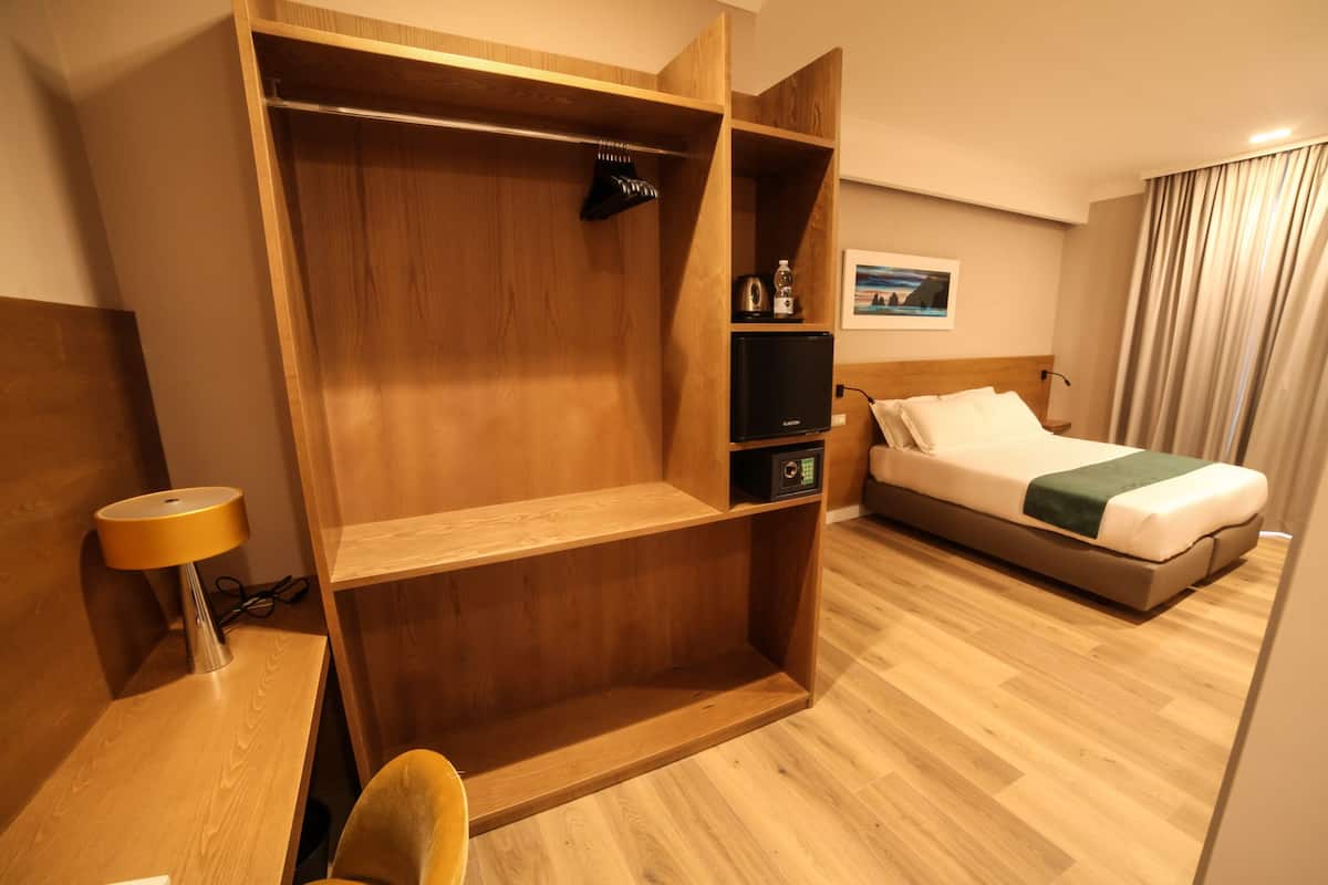 Quarto de hotel com guarda-roupa de madeira, cama com roupa de cama branca, escrivaninha com abajur e uma pequena prateleira com chaleira e garrafa.