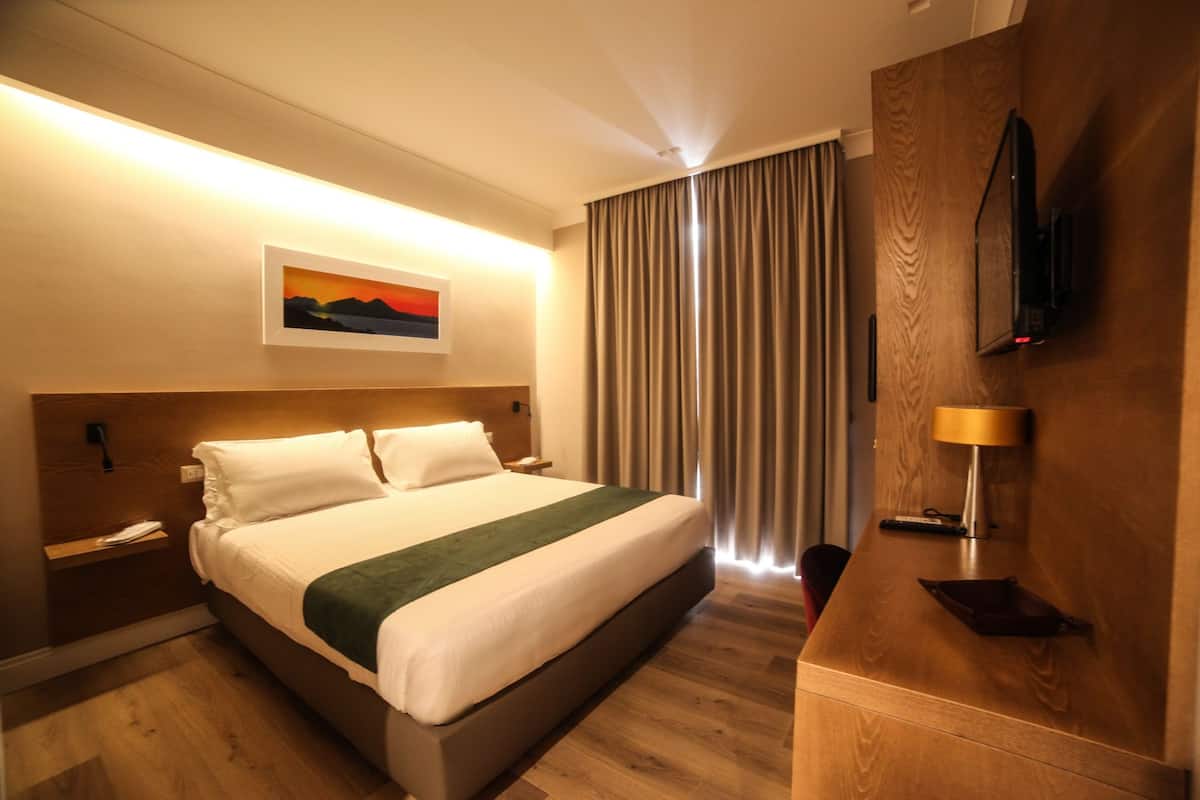 Quarto de hotel com uma cama, um quadro acima dela, uma televisão na parede e uma escrivaninha de madeira com uma luminária.