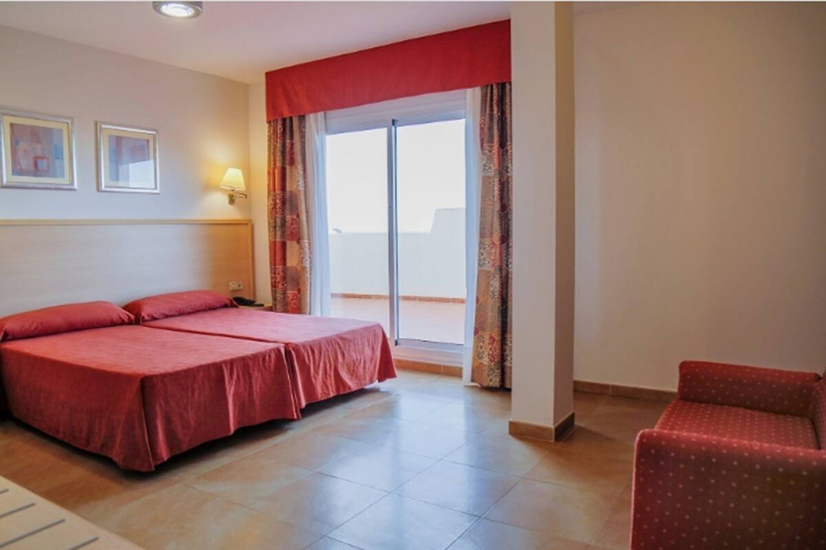 Quarto de hotel com cama, cadeira, janela com cortinas e luminária fixa na parede.