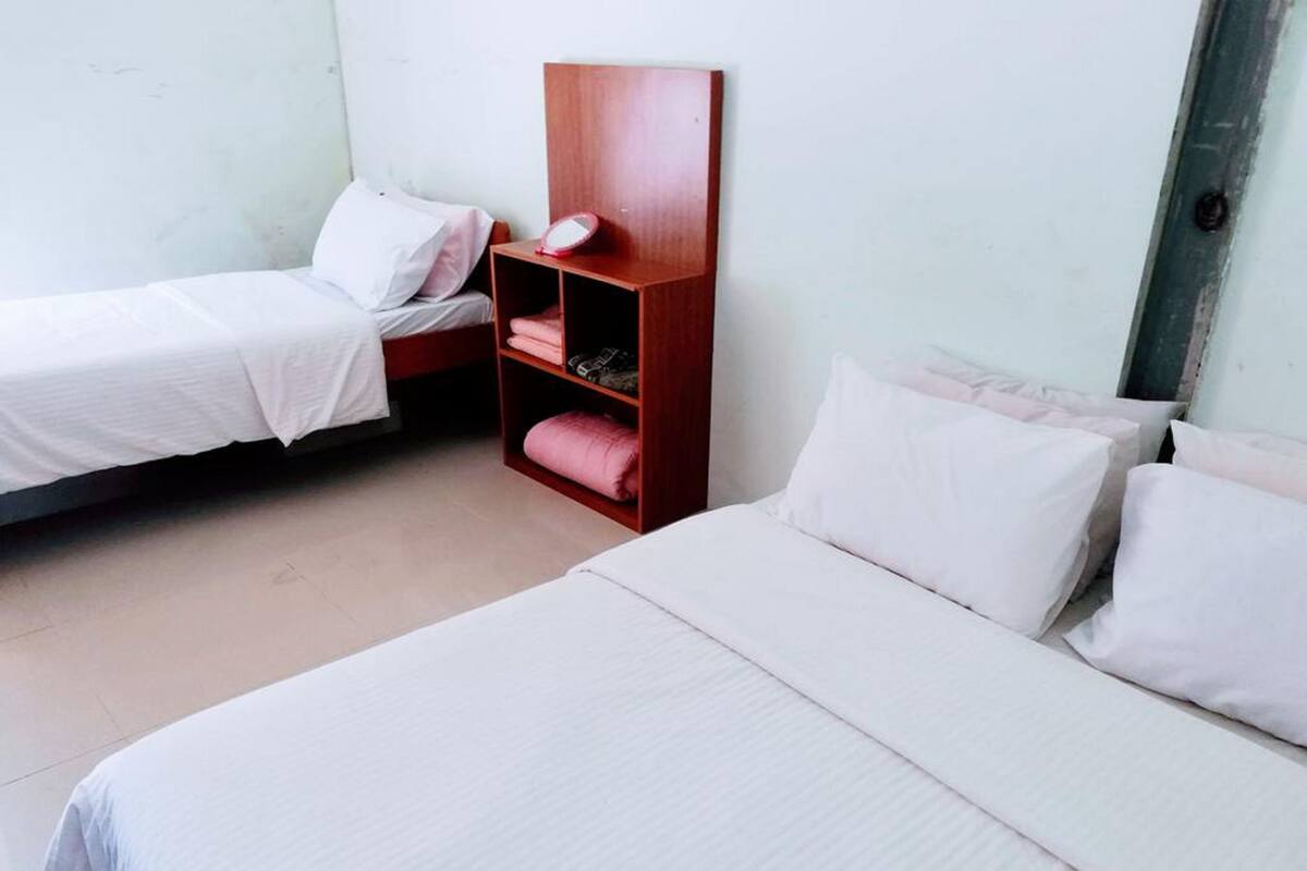 Une chambre simple et bien rangée, avec deux lits, une table de chevet en bois dotée d’un miroir et une étagère où sont pliées des serviettes.