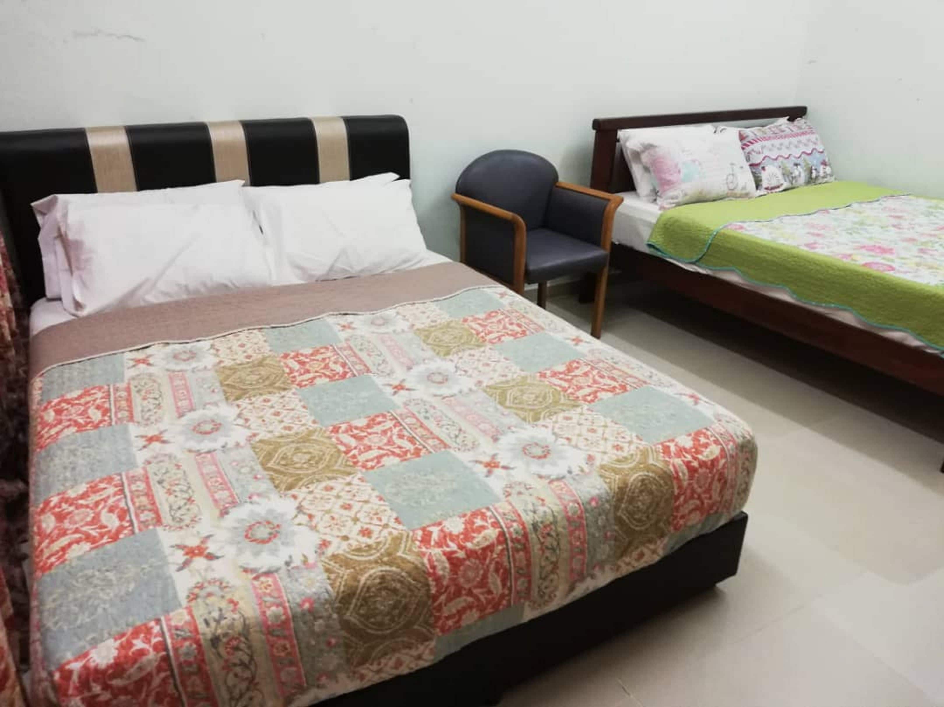 Une chambre avec deux lits, l’un recouvert d’une courtepointe à motifs et l’autre d’un couvre-lit vert. On y trouve également une chaise et une petite table.
