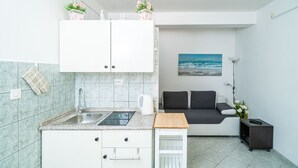 Una cucina compatta con lavello, piano cottura e pensili, una piccola zona pranzo con tavolo e sedie, e un divano con un quadro che raffigura l'oceano alle spalle.