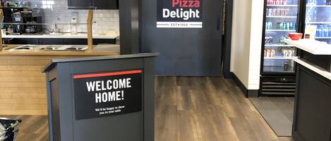 A modern pizza restaurant interior with a welcome sign, a Coca-Cola refrigerator, and a counter with a coffee machine.