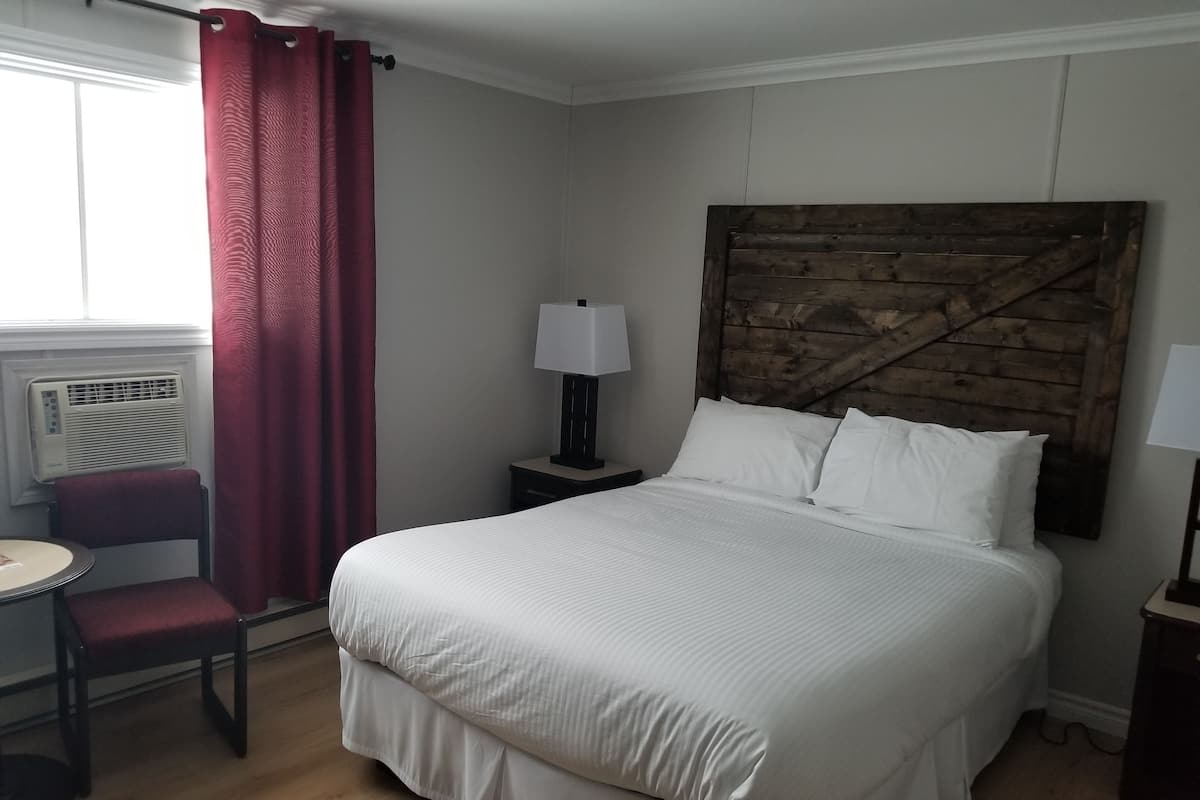 Habitación de hotel con una cama grande, cabecera de madera, mesita de noche con lámpara, una silla y una ventana con cortinas.