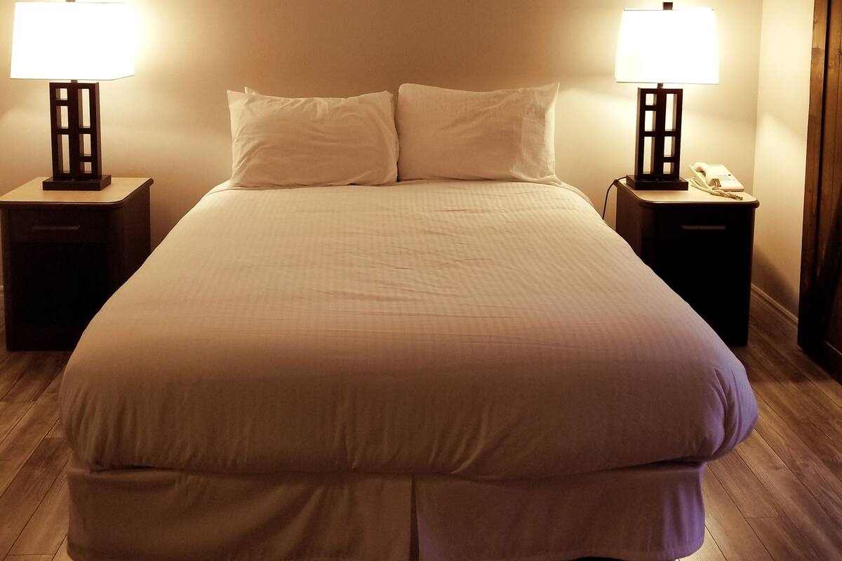 Una cama grande con edredón blanco en una habitación de hotel. Hay dos mesitas de noche, cada una con una lámpara y un teléfono. El suelo es de madera y las paredes son lisas.