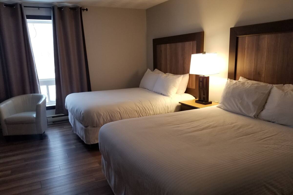 Habitación de hotel con dos camas, una silla, una mesita de noche con lámpara y una ventana con cortinas.