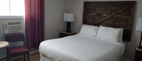 A hotel room with a large bed, a wooden headboard, a small table with a chair, a lamp, and a window with curtains.
