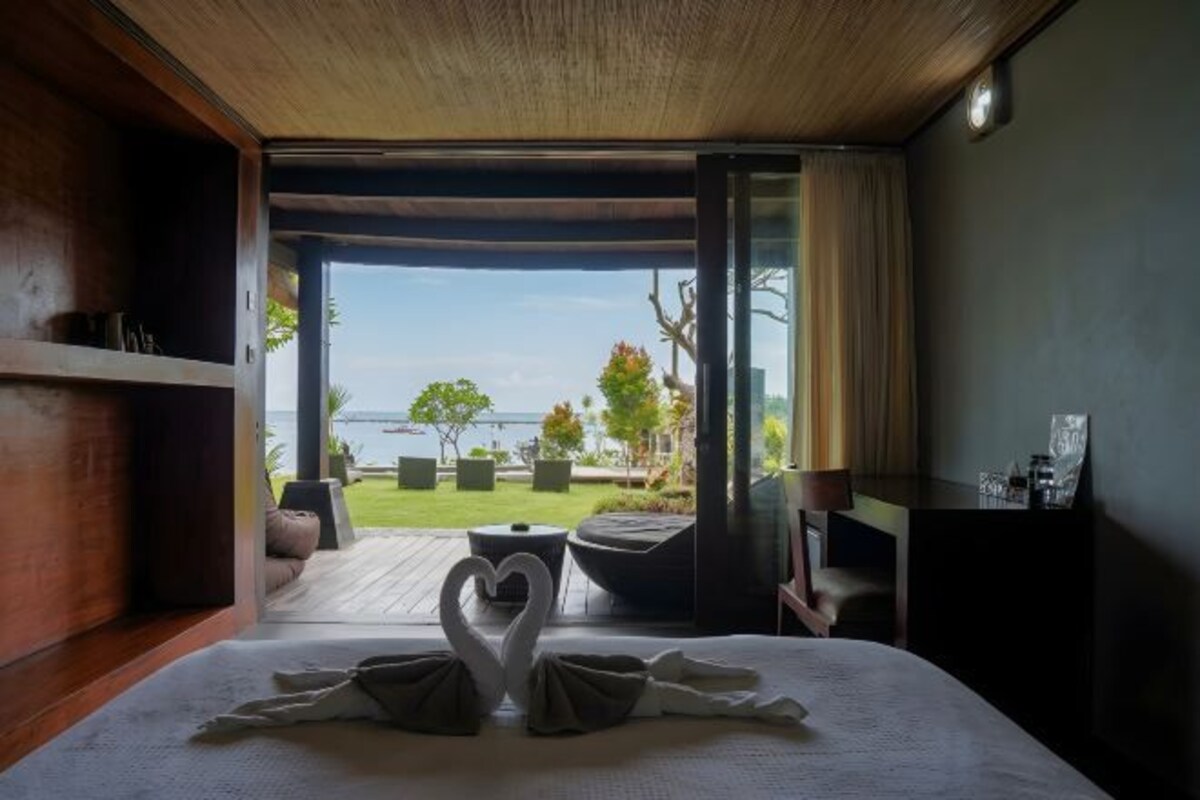 A hotel room with a bed, a heart-shaped towel arrangement, a view of the sea, and a wooden ceiling.