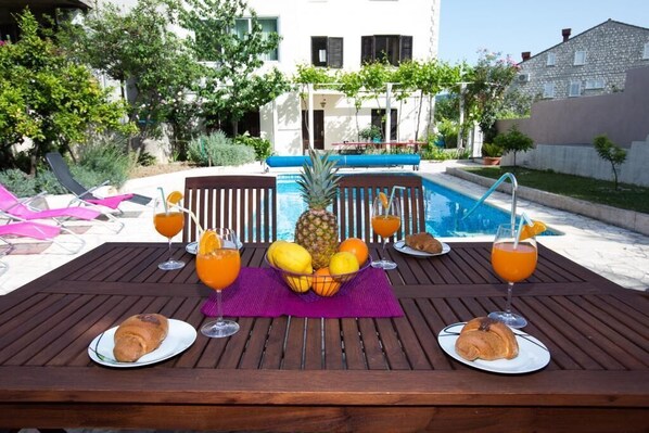 A wooden table with breakfast items and drinks, a pool, and a building in the background.