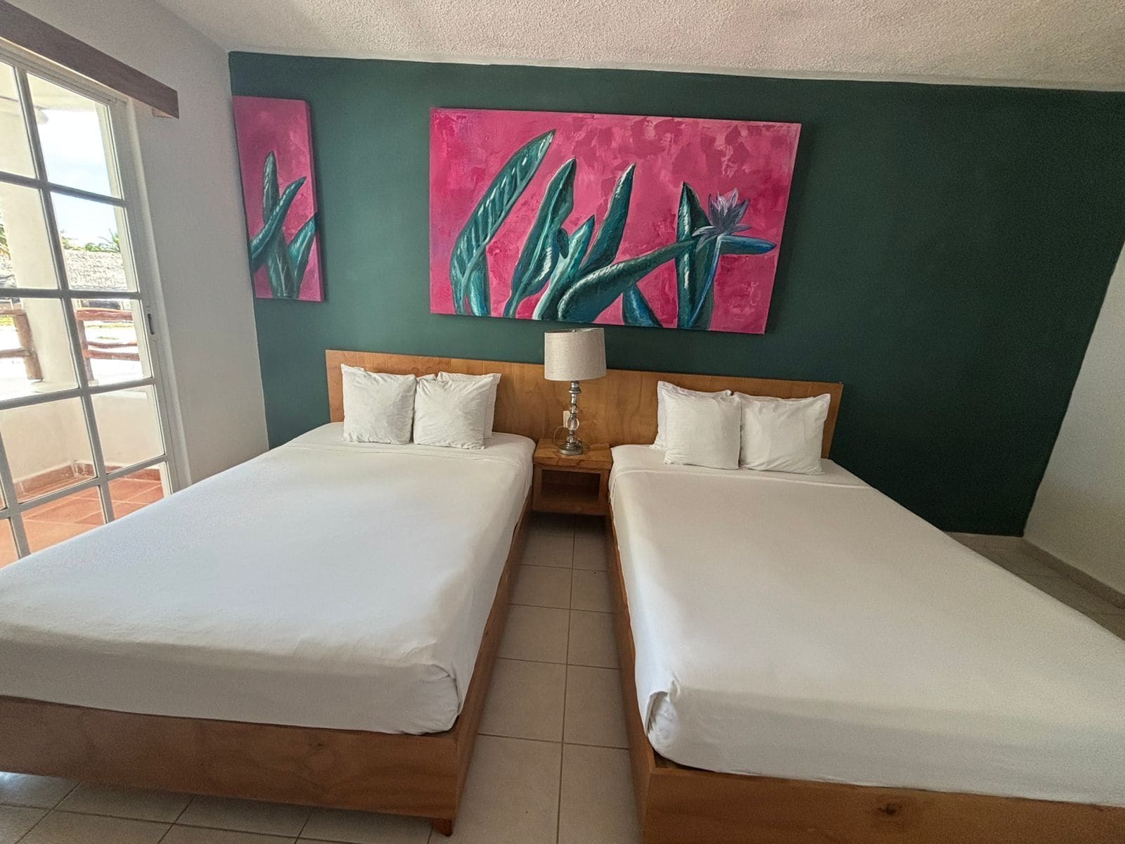 Una habitación de hotel con dos camas, cabecera de madera, mesita de noche con lámpara y dos cuadros grandes en la pared.