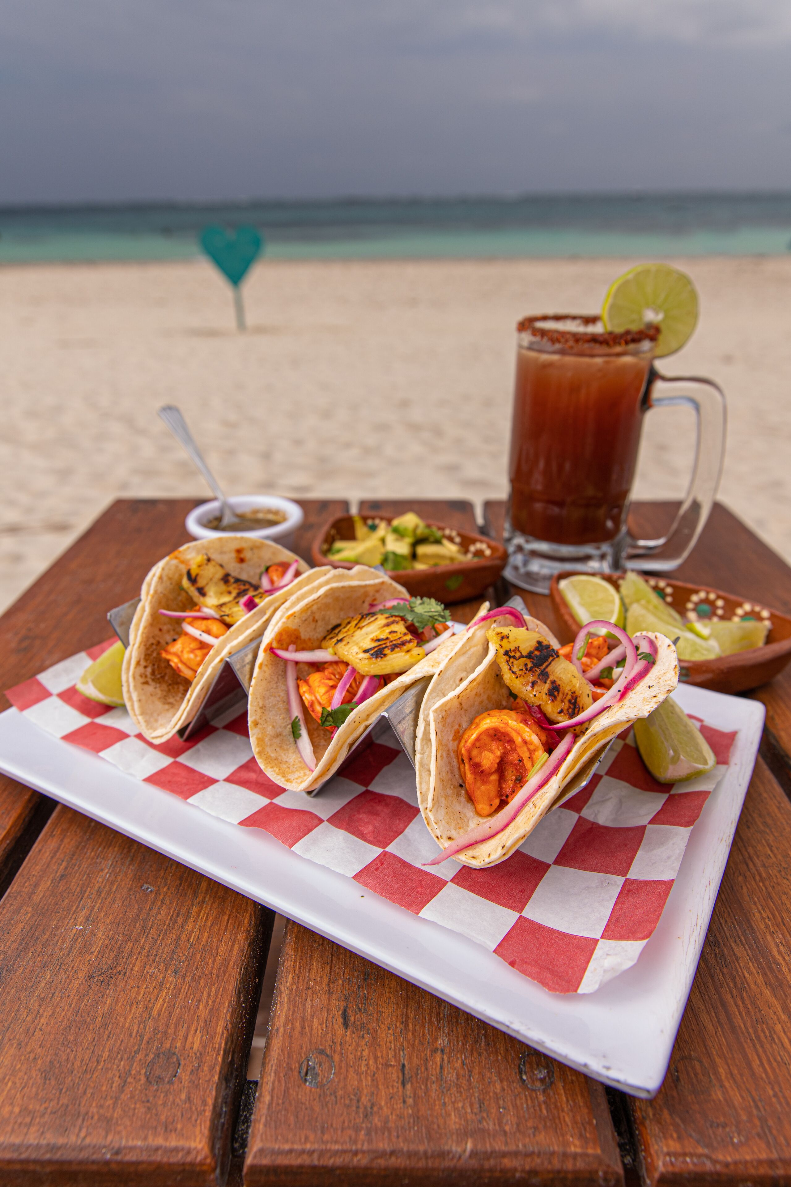 Three tacos on a wooden table with a beach view in the background.
