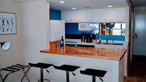 A compact kitchen with white cabinets, a blue backsplash, a microwave, and a wooden countertop. There are three black bar stools in front of the counter.