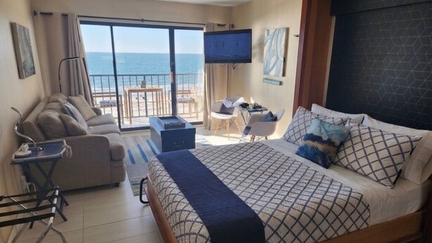 A hotel room with a bed, a sofa, a desk, a TV, and a balcony with a view of the sea.