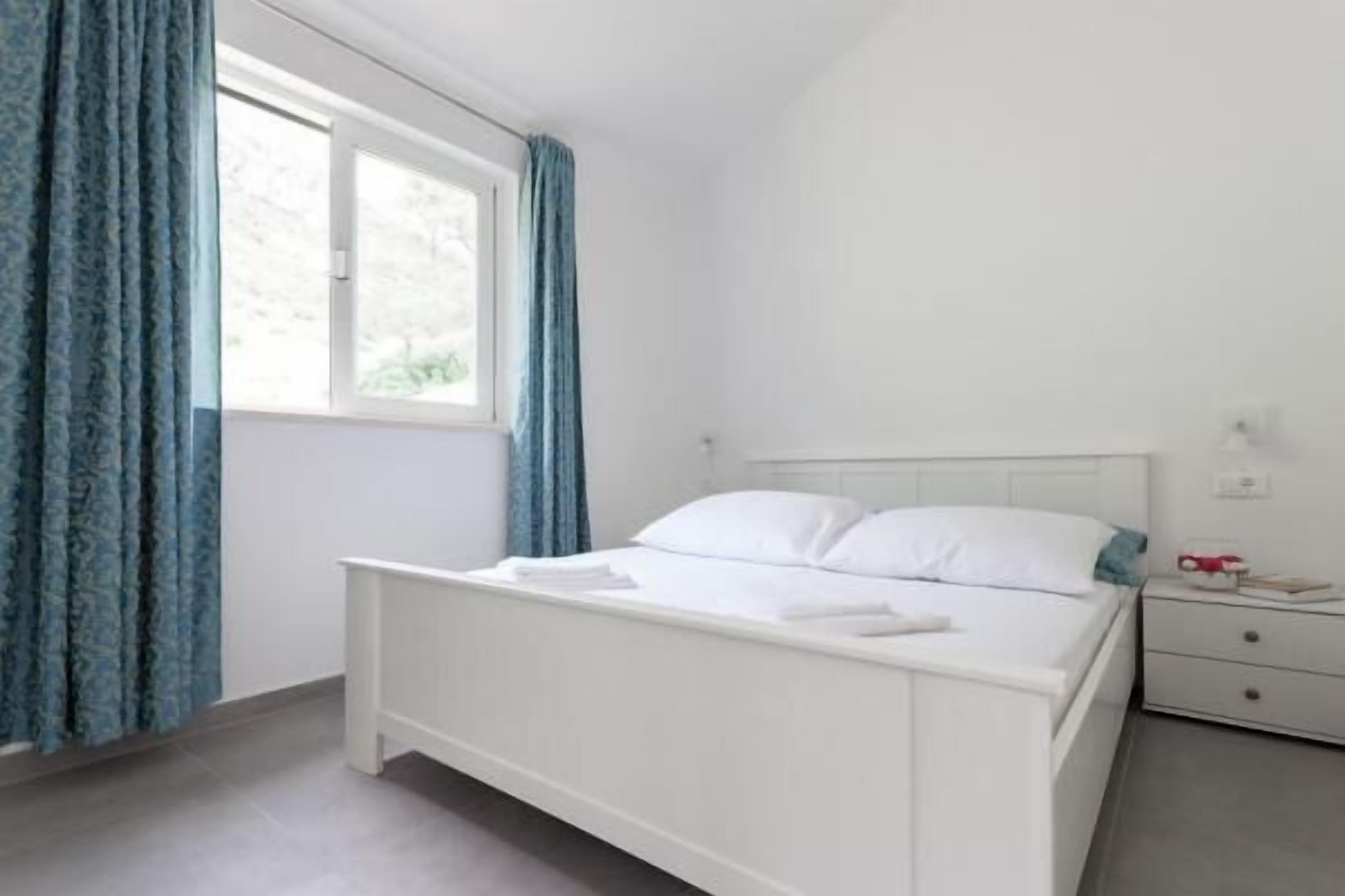 Een wit bed met hoofdeinde, een nachtkastje met lamp en een raam met gordijnen.