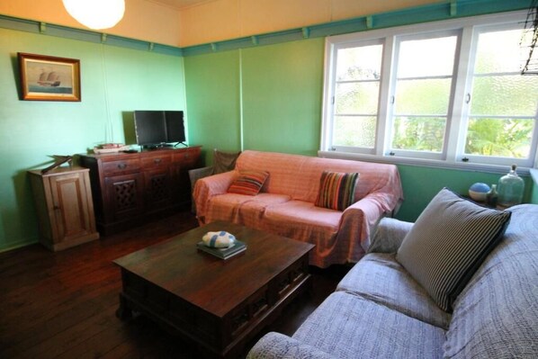 Una sala de estar con una pared verde, una ventana con marcos blancos, un mueble de madera, un televisor, un sofá rosa y un sofá gris, junto con una mesa de centro de madera.