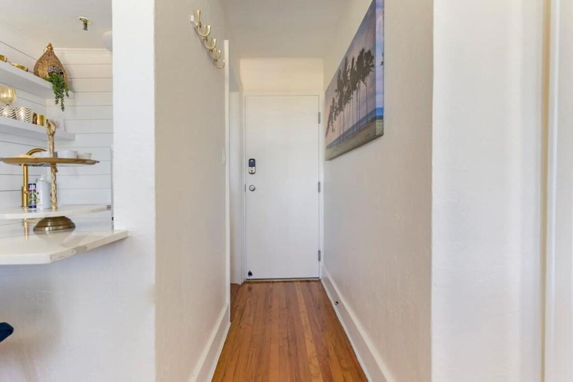 A hallway with a wooden floor, white walls, a door, and a shelf with decorative items.