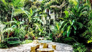 A garden patio with a round table and benches, surrounded by lush greenery. - Pool/StepsToBeach/Dining Delight 1br (Palm Beach)