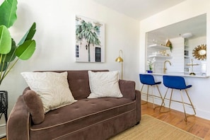 Una sala de estar con un sofá marrón, dos sillas azules, una planta verde grande y una cocina con gabinetes blancos y un grifo dorado.