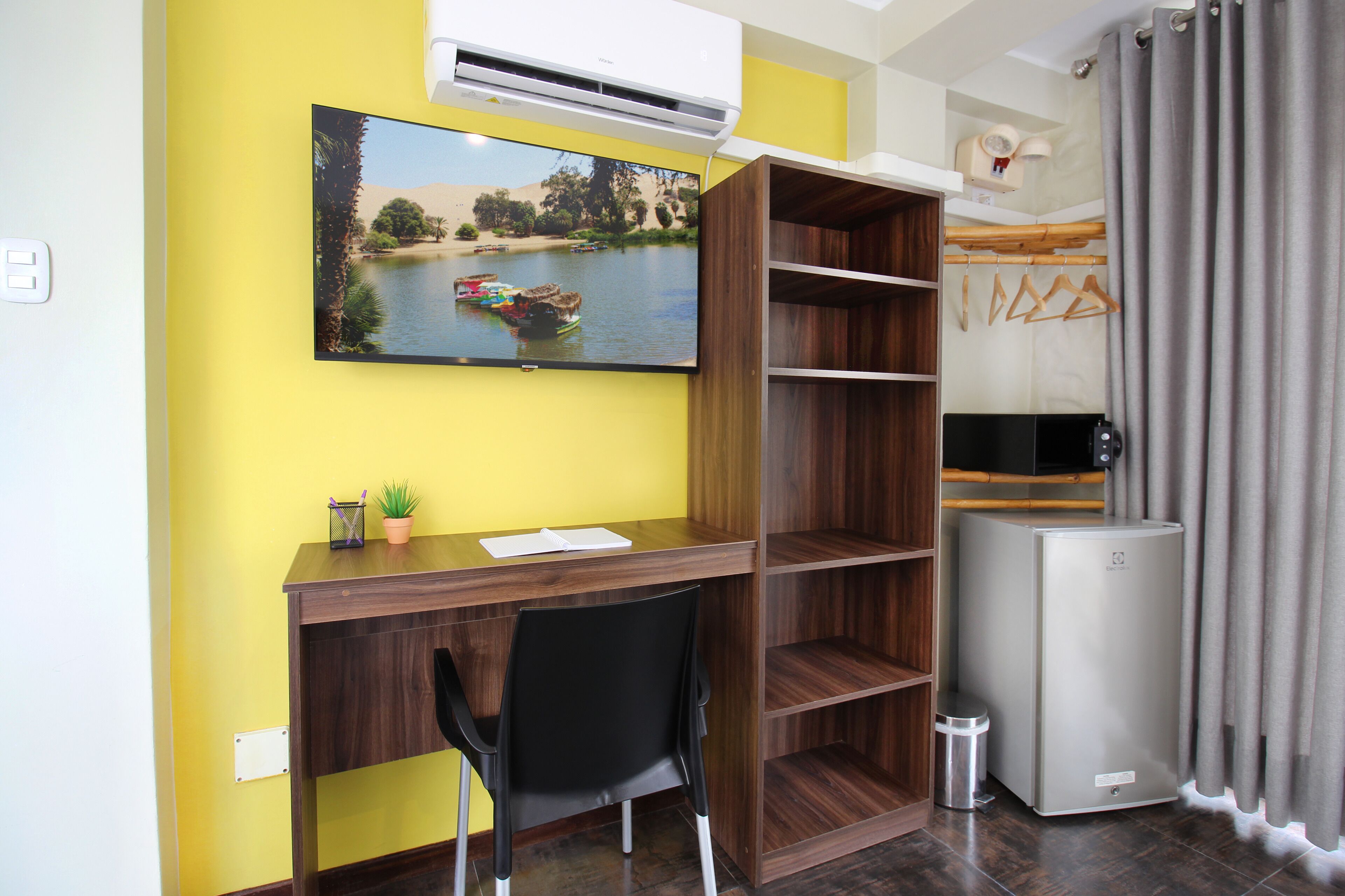 Een kleine kamer met een houten bureau, een zwarte stoel, een aan de muur bevestigde televisie en een kleine koelkast.
