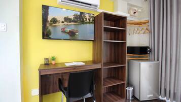 Una habitación compacta con un escritorio de madera, una silla negra, un televisor montado en la pared y un refrigerador pequeño.
