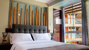 A hotel room with a large bed, a headboard with vertical bamboo elements, a view of the outside balcony, and a wooden door. - Bananas Adventure Hostel (Ica)