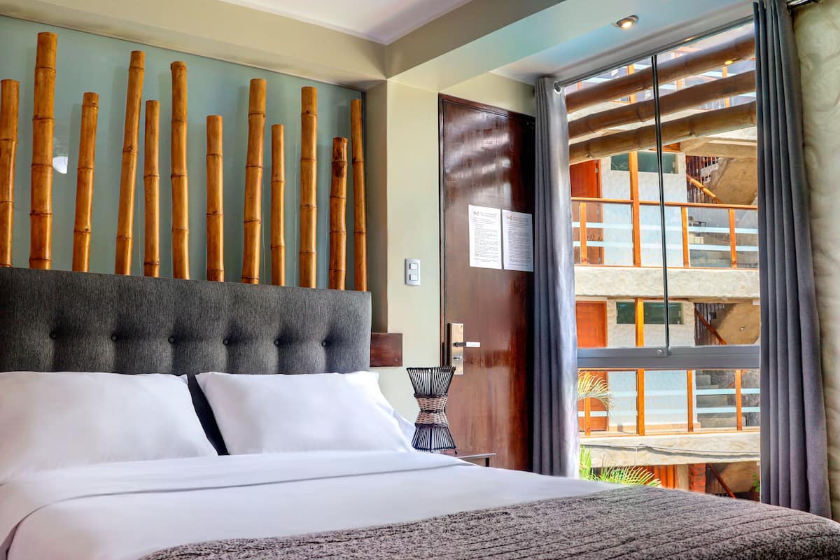 Habitación de hotel con una cama grande, cabecera con elementos verticales de bambú, vista al balcón exterior y una puerta de madera.