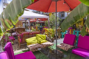 An outdoor seating area with colorful chairs and tables, surrounded by potted plants and a large umbrella. - Bananas Adventure Hostel (Ica)