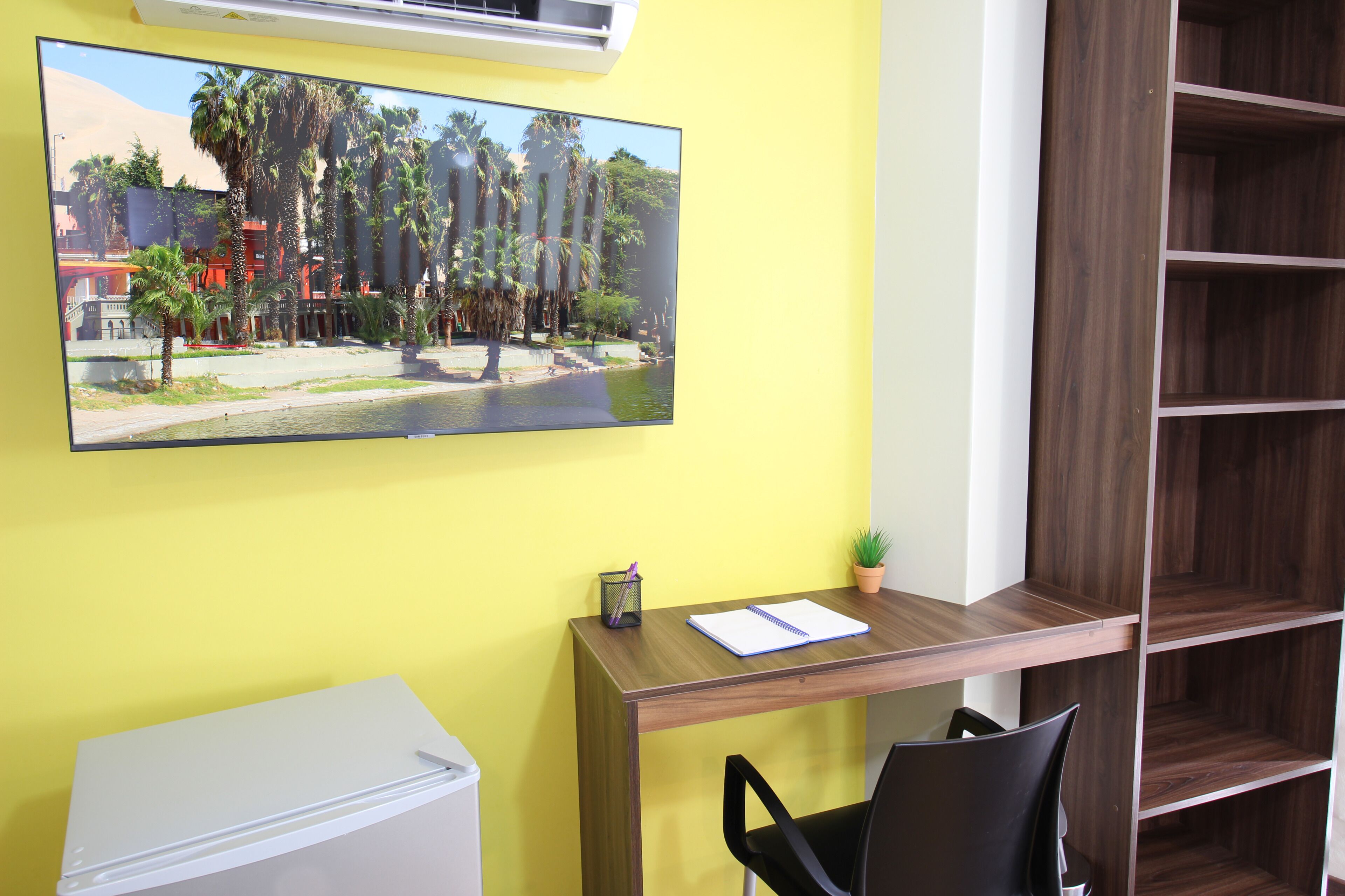A room with a wooden desk, a chair, a television mounted on the wall, and a small potted plant.