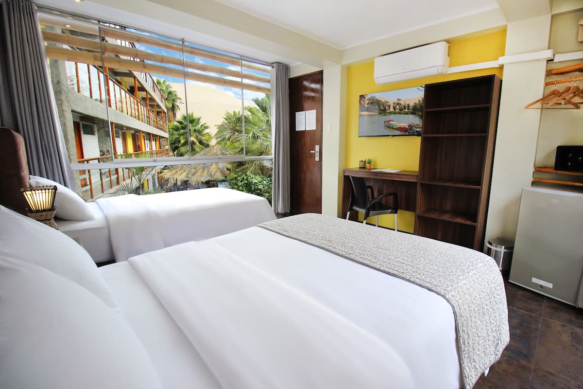 Habitación de hotel con dos camas, un escritorio, una silla, un armario y vistas a un edificio con balcones y palmeras.