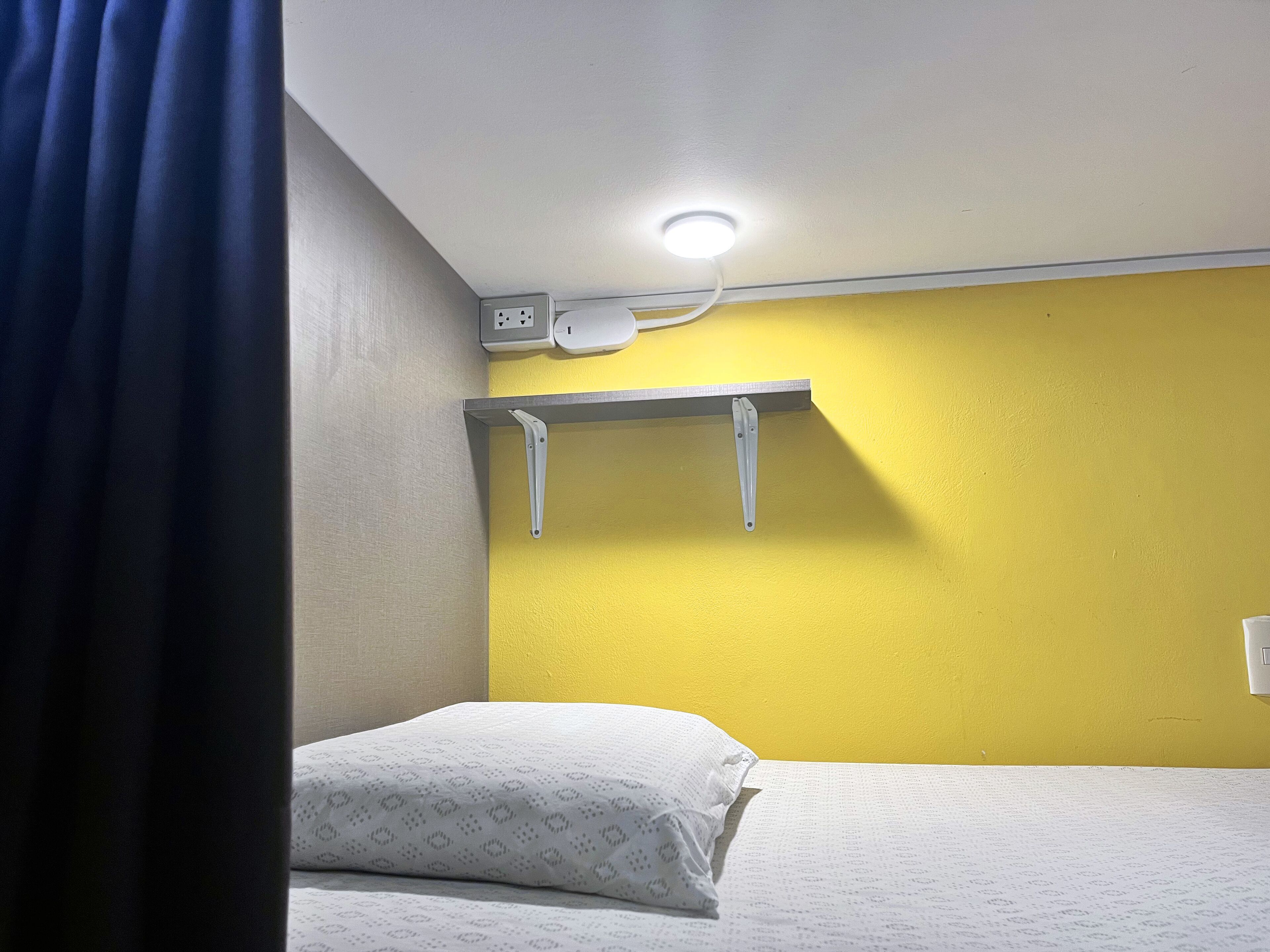 A small, single-bed room with a yellow wall, a white ceiling light, and a power outlet above the bed.