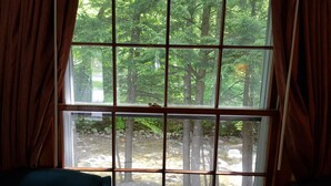 A room with a large window divided into nine panes, offering a view of a forested area with trees and a rocky stream. - Franconia Notch Motel (Lincoln)
