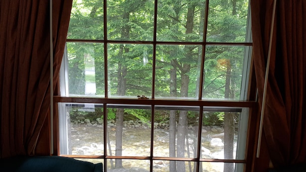 A room with a large window divided into nine panes, offering a view of a forested area with trees and a rocky stream. - Franconia Notch Motel (Lincoln)